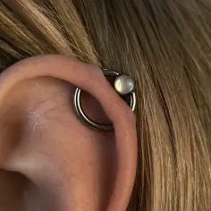 Oryginalny kolczyk orbital piercing z białym kamieniem ozdabia ucho, otoczony blond włosami.