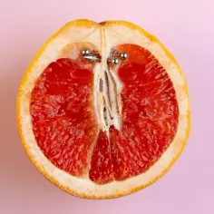 Przekrojony grejpfrut z metalowymi ozdobami, przypominającymi guiche piercing, na różowym tle.