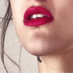 Usta z czerwoną szminką i subtelnym ashley piercingiem. Widać też tatuaż na szyi.