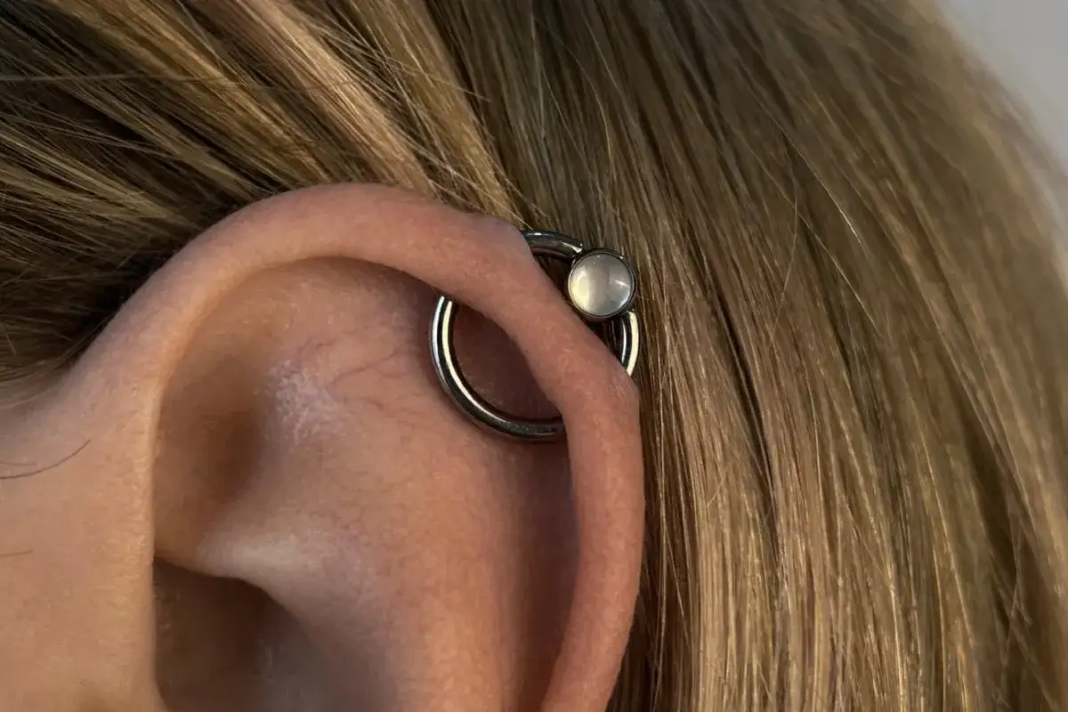 Oryginalny kolczyk orbital piercing z białym kamieniem na uchu, otoczony blond włosami.